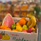 A cardboard box filled with assorted fruits, including a watermelon slice, oranges, bananas, and apples, set in a blurred indoor environment.