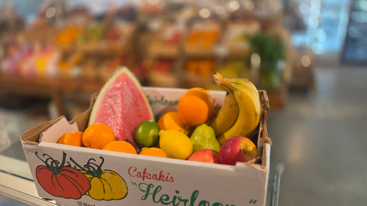 A cardboard box filled with assorted fruits, including a watermelon slice, oranges, bananas, and apples, set in a blurred indoor environment.